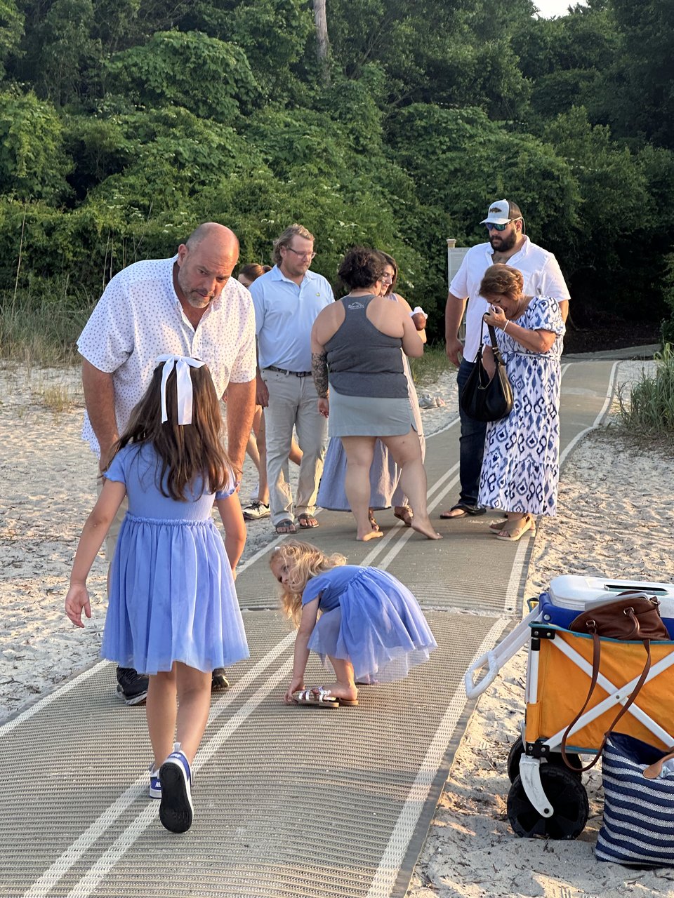 Adults and children gather on a sandy beach path in warm evening light.