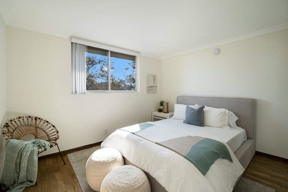 Staged bedroom with a gray upholstered bed, teal accents, woven chair, and window overlooking trees.