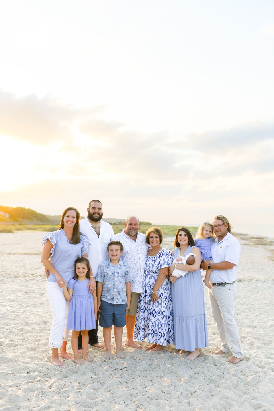 Family of ten posing on a beach at golden hour, including children and a newborn.