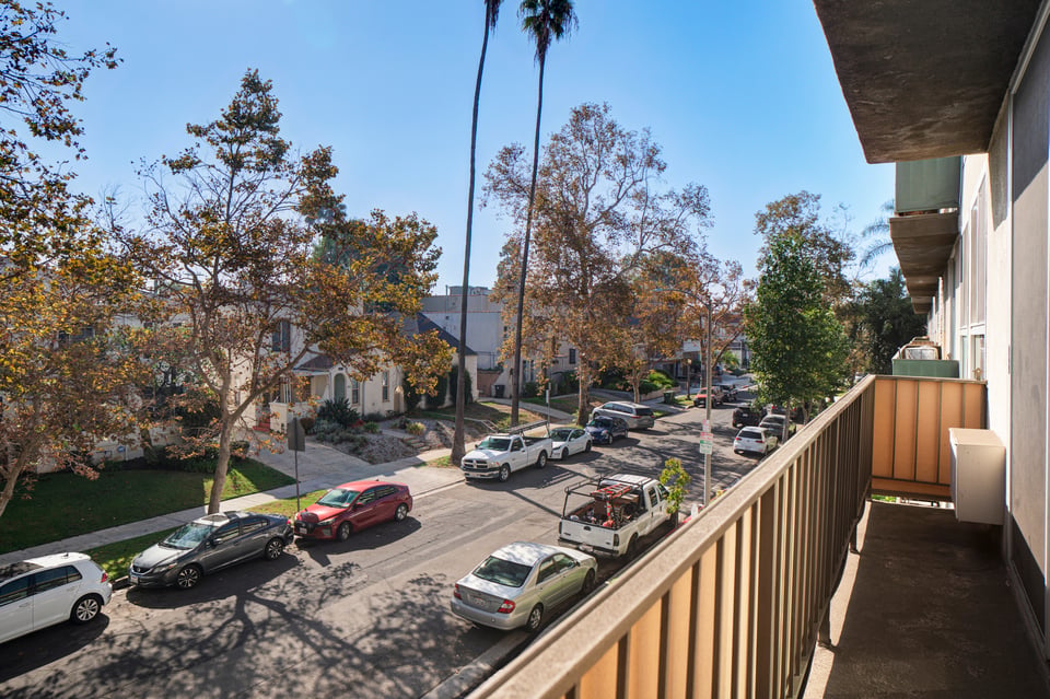 Apartment balcony overlooking a tree-lined residential street with parked cars.