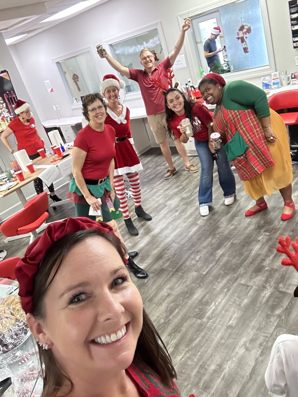 Seven adults in festive holiday outfits pose for a group selfie at an indoor event.