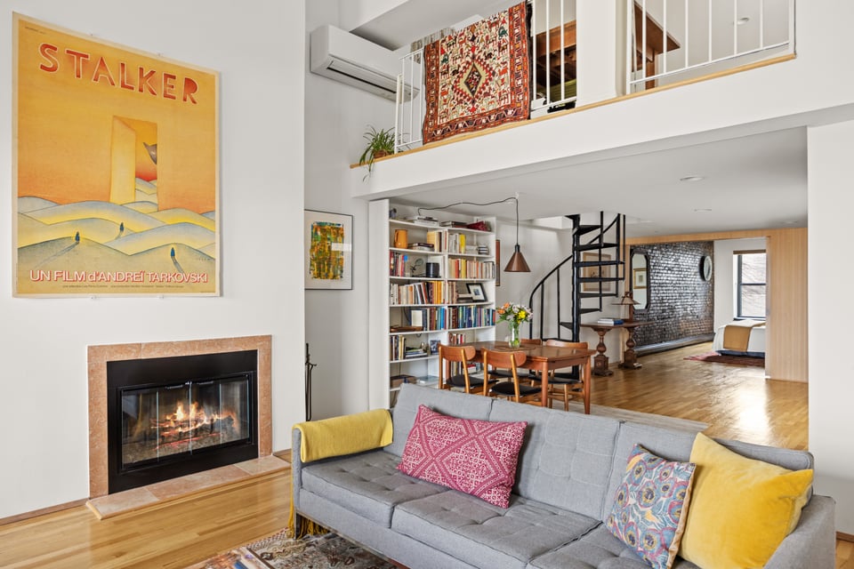 Open-plan living room with a fireplace, colorful sofa, bookshelves, and spiral staircase to a mezzanine.