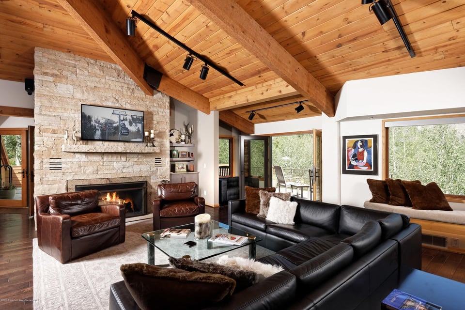 Living room with a stone fireplace, vaulted wood ceiling, leather seating, and large windows overlooking trees.