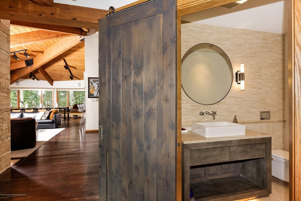 Bathroom vanity with a vessel sink and round mirror; an open barn door reveals a timber-beamed living room.