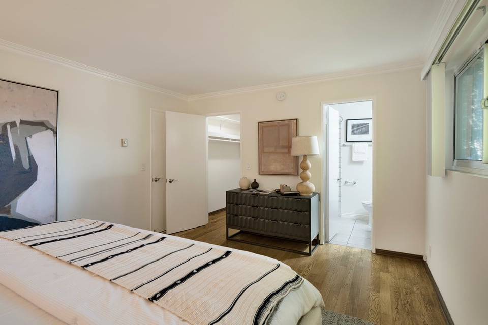 Bedroom with a striped white quilt, dark dresser, hardwood floors, and an adjoining bathroom.