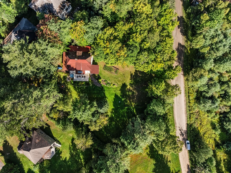 Aerial view of a red-roofed house surrounded by dense trees and lawn beside a rural road.