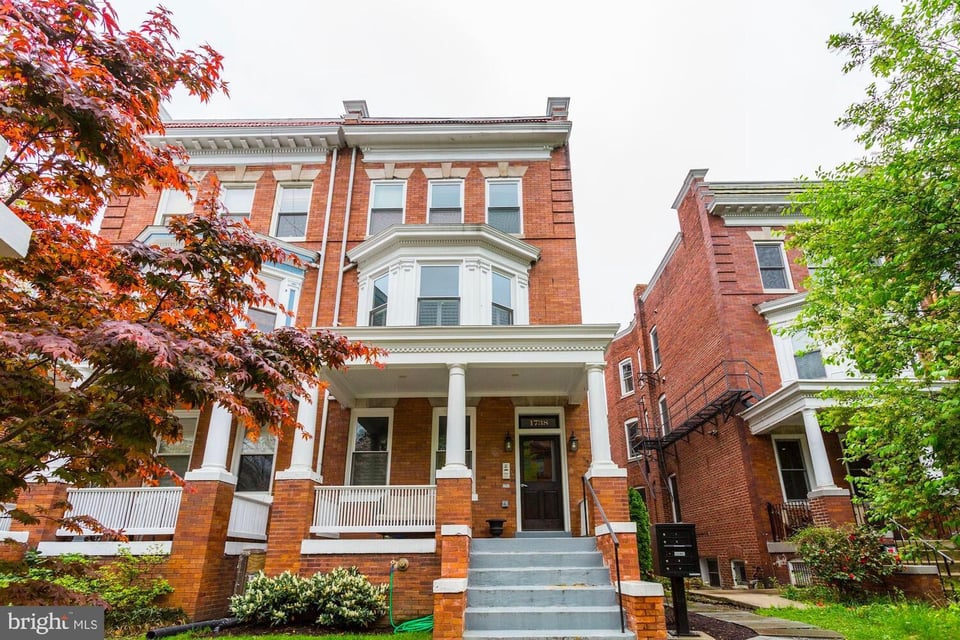 Red-brick rowhouse with a columned porch and broad front steps, framed by leafy trees.