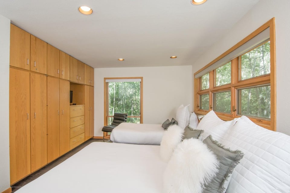 Bedroom with two white beds, extensive built-in wood cabinetry, and large windows overlooking trees.