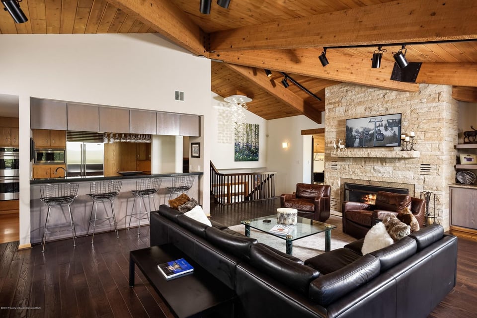 Open-plan living room and kitchen with vaulted wood ceiling, leather seating, and a stone fireplace.