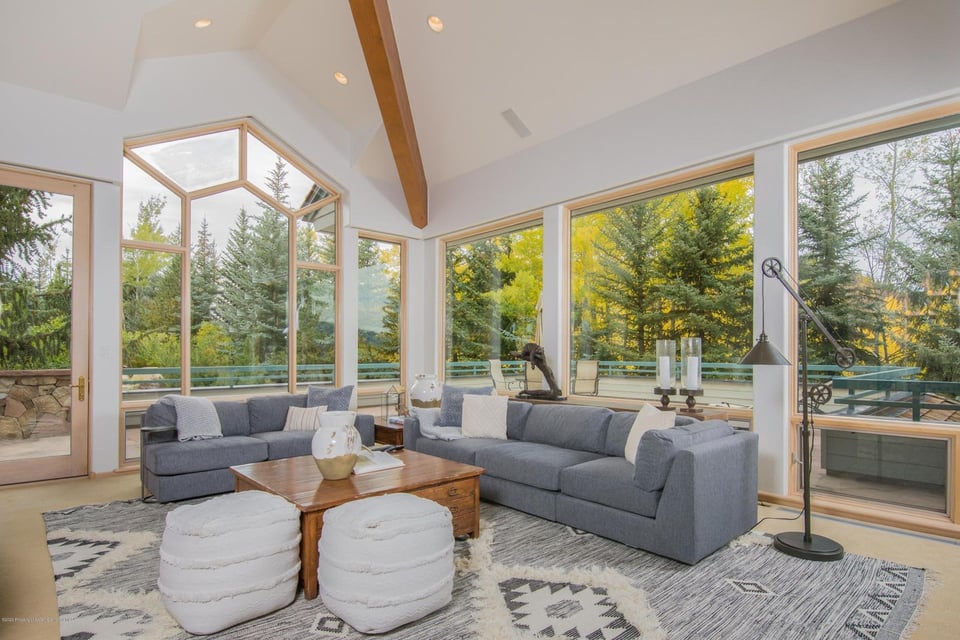 Living room with gray sofas, white ottomans, and tall windows overlooking trees.