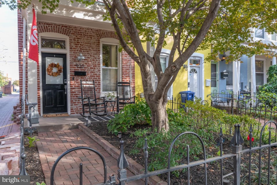 Red-brick rowhouse with a black front door, rocking chairs, and a leafy tree shading its fenced garden.