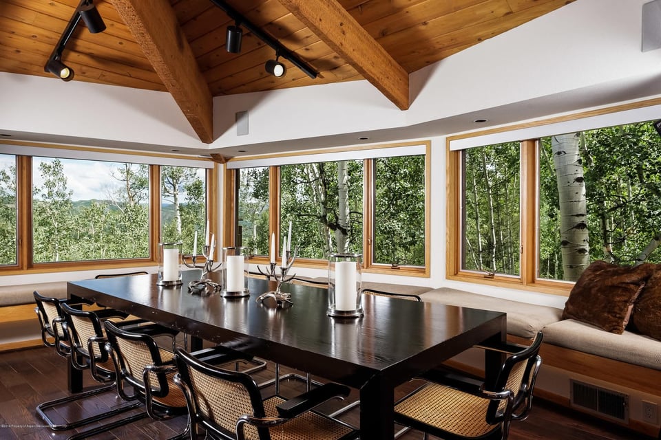 Dining room with a long dark table, vaulted wood ceiling, and windows overlooking trees and mountains.