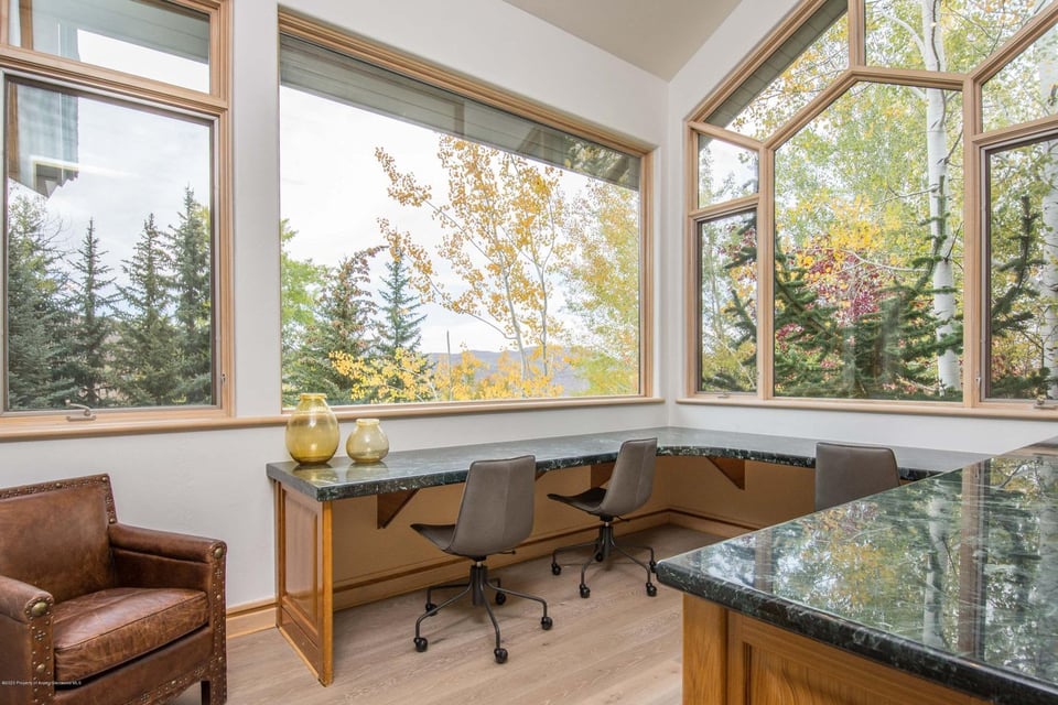 Home office with wraparound counters, leather armchair, and large windows overlooking autumn trees and mountains.