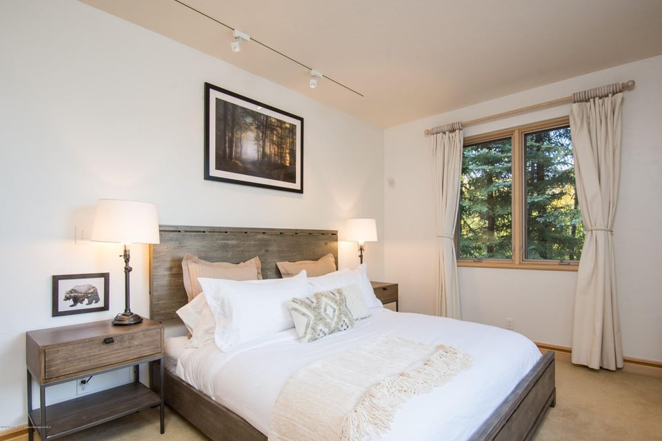 Bedroom with white bedding, rustic wood furniture, and a window overlooking evergreen trees.