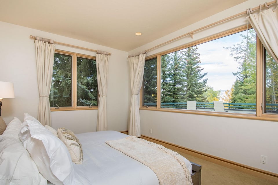 Bedroom with a white bed and large windows overlooking evergreen trees and a deck.