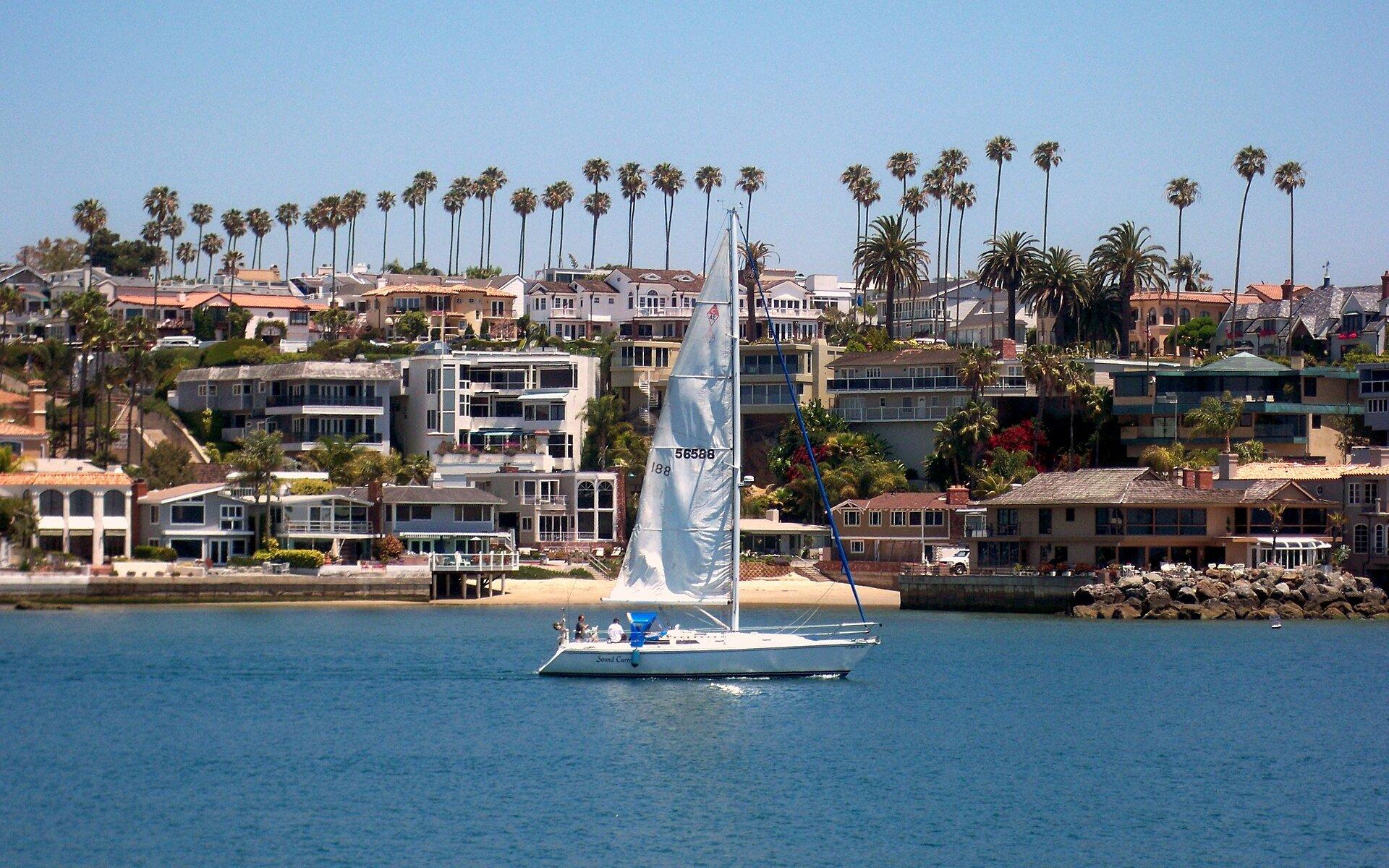 Sailboat passing waterfront homes in Newport Harbor, Newport Beach