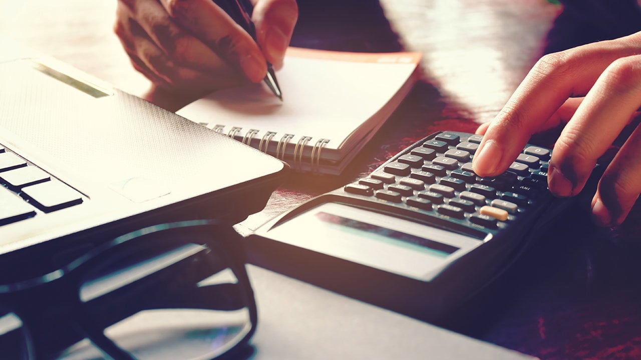 Hands write in a notebook and use a calculator beside a laptop and eyeglasses.