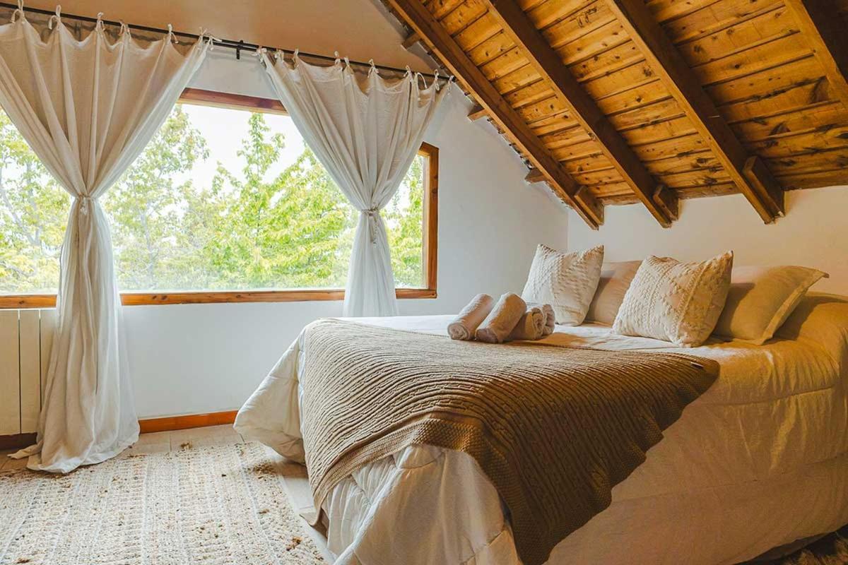 Bedroom with a large neutral bed beneath a sloped timber ceiling and a curtained window.