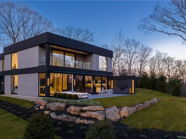 Modern glass-fronted home at dusk, with a stone retaining wall and patio.