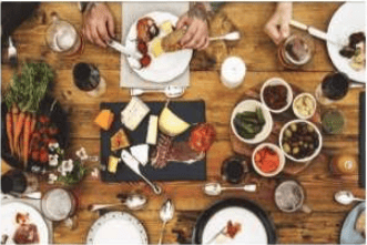 Shared meal spread across a wooden table with cheeses, meats, vegetables, and small bowls of dips.