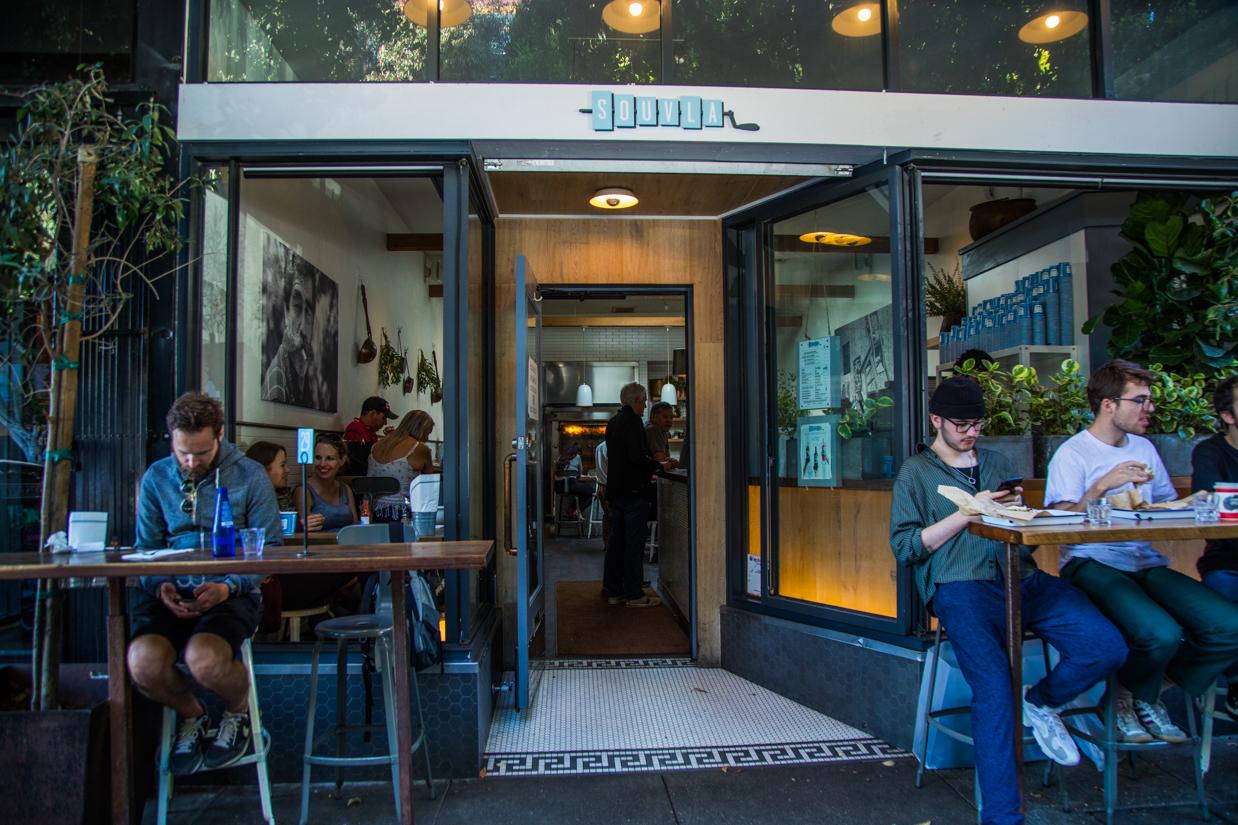 Souvla, a casual dining spot in San Francisco, showcasing an open storefront with outdoor seating, patrons enjoying meals, and a cozy interior featuring modern decor and greenery.