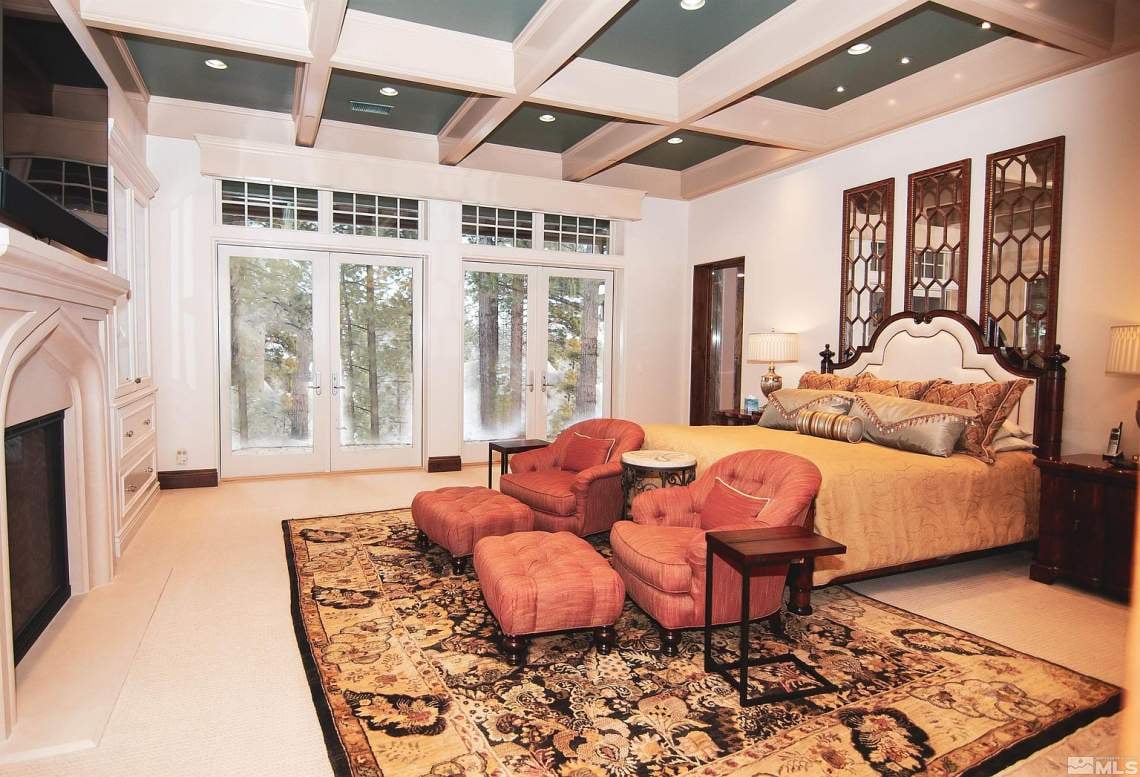 Furnished bedroom with a coffered ceiling, fireplace, and glass doors opening to a wooded view.