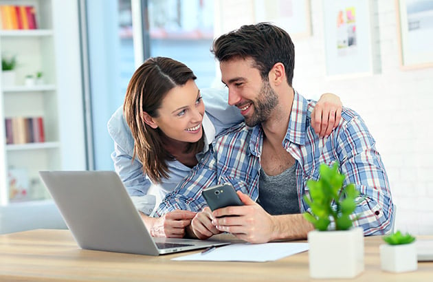 Couple smiling over a phone beside an open laptop at a table.