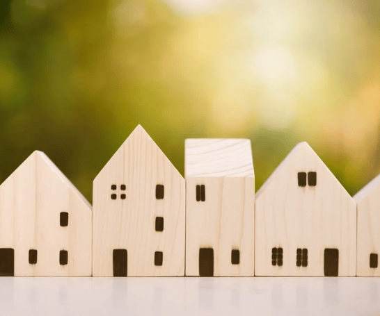 Pale wooden house models lined up against a softly blurred green and gold background.