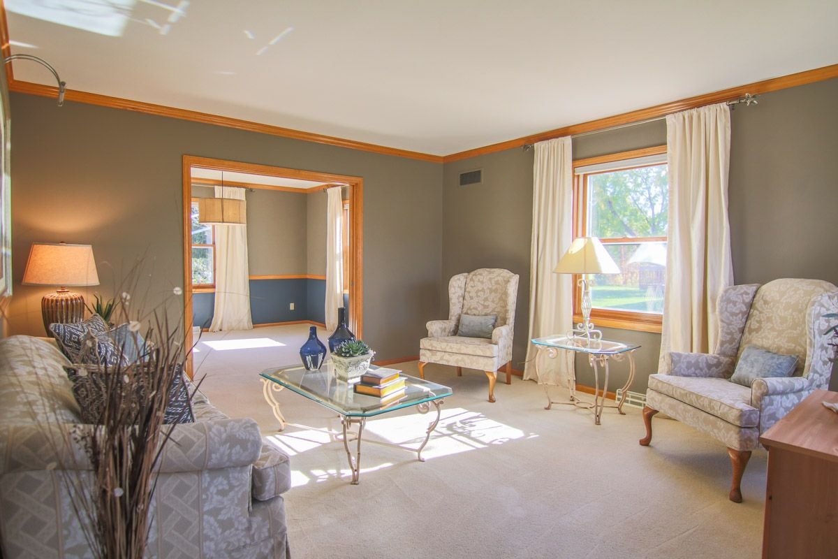 Furnished living room with patterned armchairs, a glass coffee table, and sunlight through tall windows.