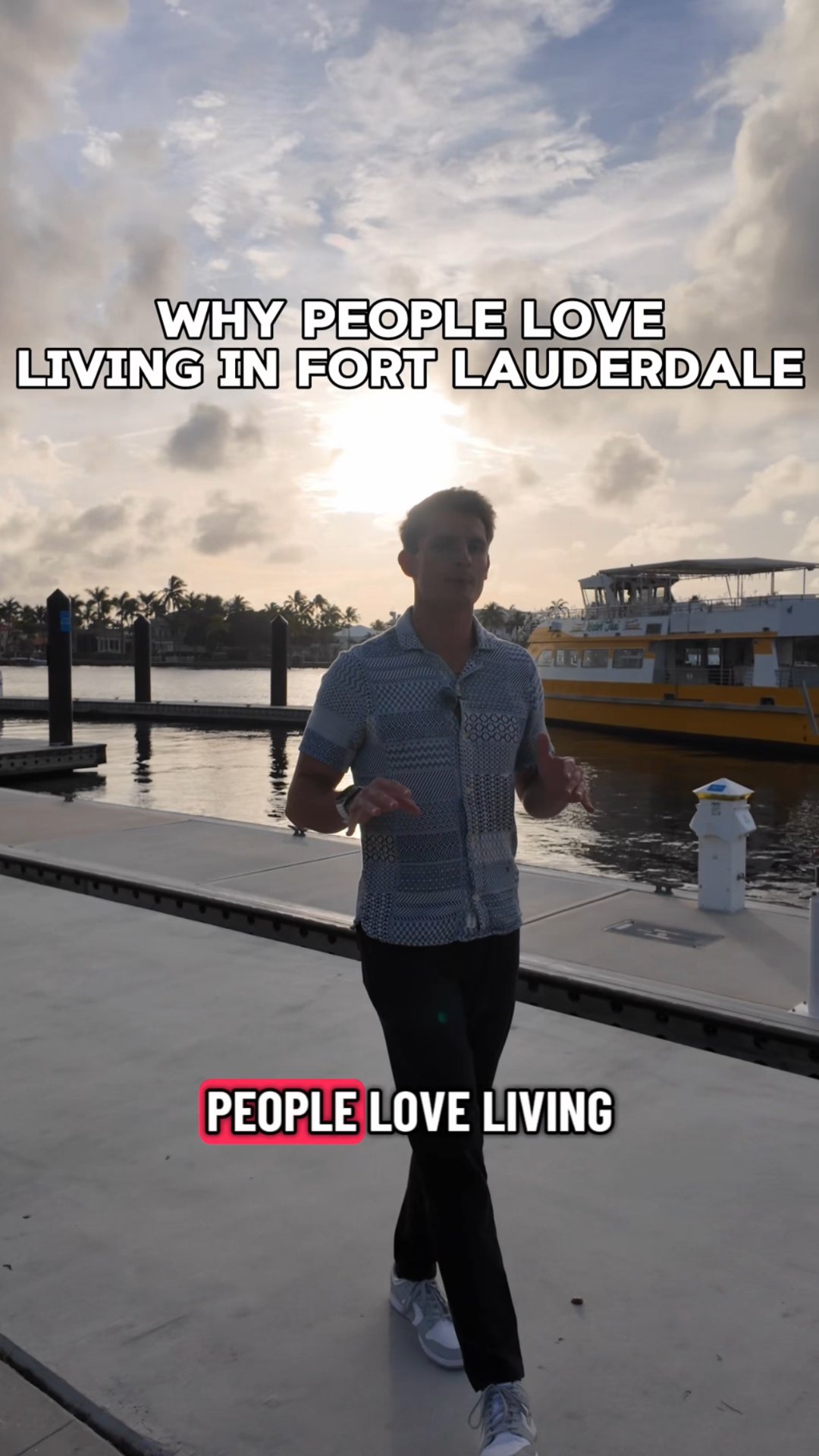 A man gestures on a waterfront dock at sunset, with a passenger boat and palm-lined shore behind him.