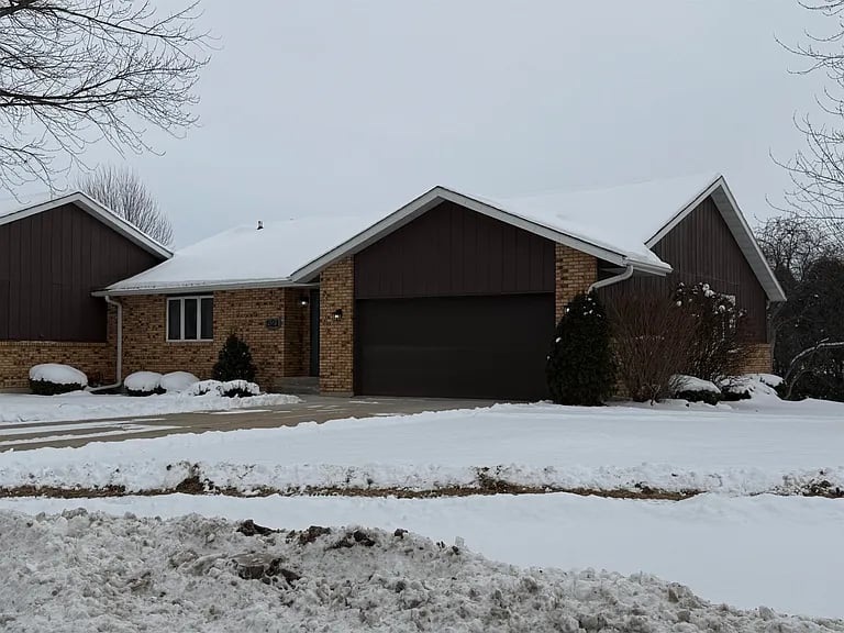 Brick home with a dark attached garage and snow-covered roof, lawn, and driveway.