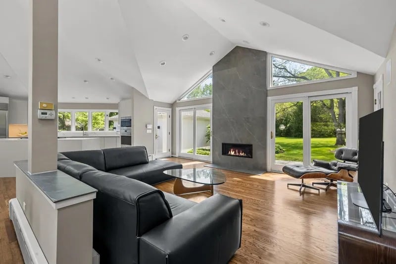 Vaulted living room with a stone fireplace, black leather seating, and windows overlooking a green garden.