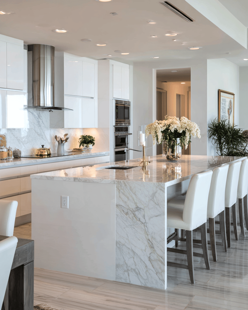 Modern white kitchen with a veined marble island, gold faucet, and white flowers.