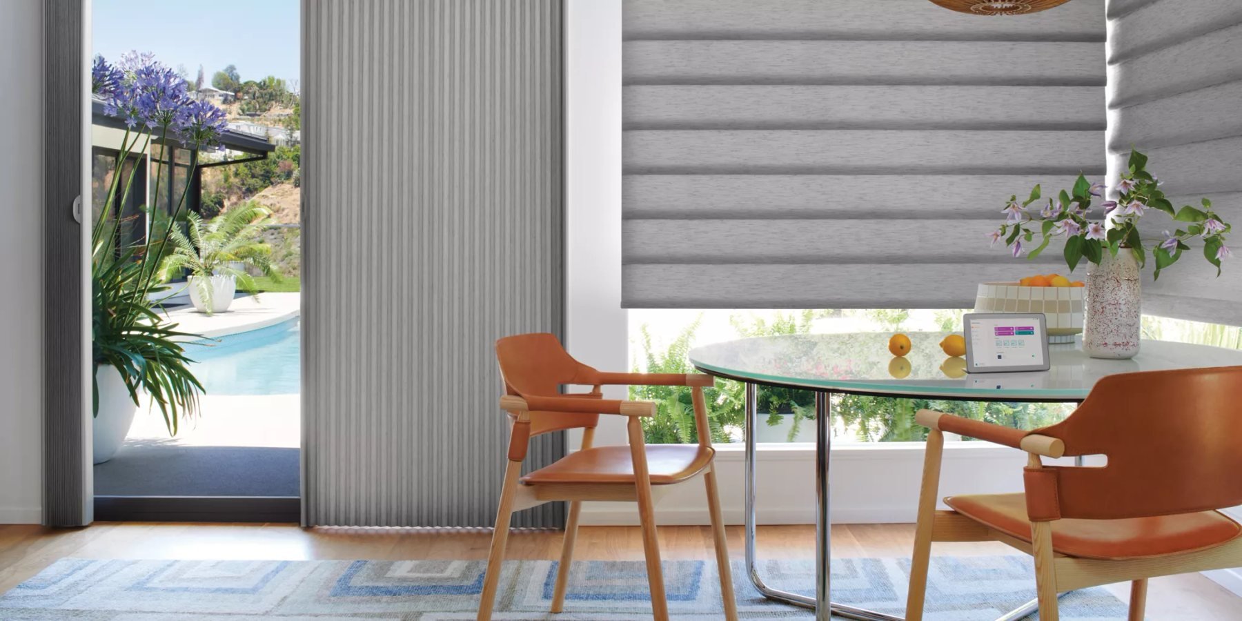 A dining nook with orange leather chairs and a round glass table. A gray vertical cellular shade covers a sliding glass door that opens onto a pool, and a gray layered Roman shade is half raised over a wide window.