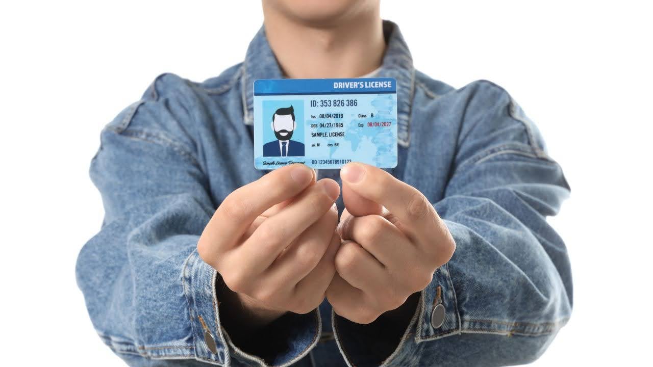 A person in a denim jacket holds up a driver's license with both hands. The card features a photo, ID number, and details, set against a white background.
