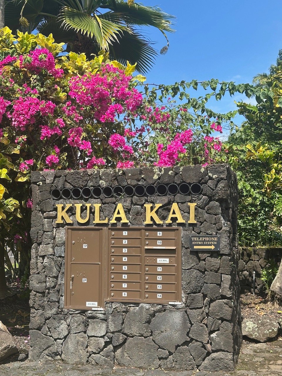 Kula Kai community sign and mailboxes beneath bougainvillea and palm fronds.
