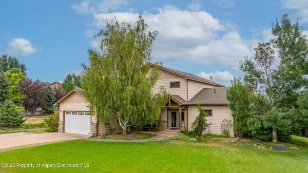 Two-story beige house with an attached garage, broad green lawn, and mature trees.