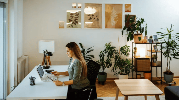 Woman working at a laptop in a bright home office surrounded by houseplants.
