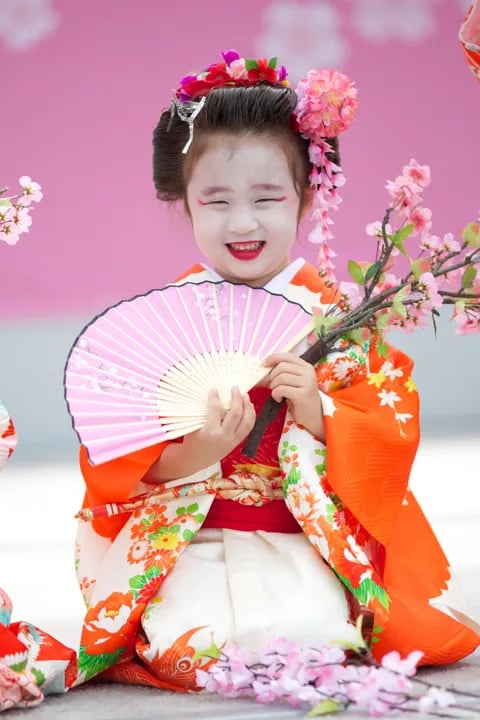 A smiling young girl in traditional makeup and an orange floral kimono holds a pink fan and cherry blossom branch.