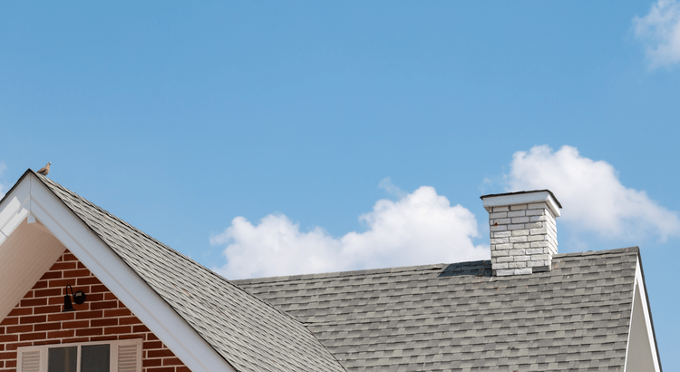 Gabled roof of a brick house with gray shingles and a white chimney under a clear blue sky with a few clouds. A small bird perches on the rooftop.