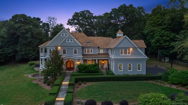 Gray two-story home with illuminated windows, a long front walkway, landscaped lawn, and mature trees at dusk.