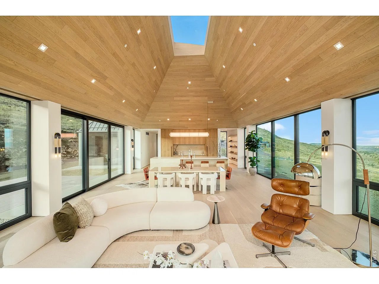 Open-plan living room with vaulted wood ceiling, skylight, and floor-to-ceiling windows overlooking hills.