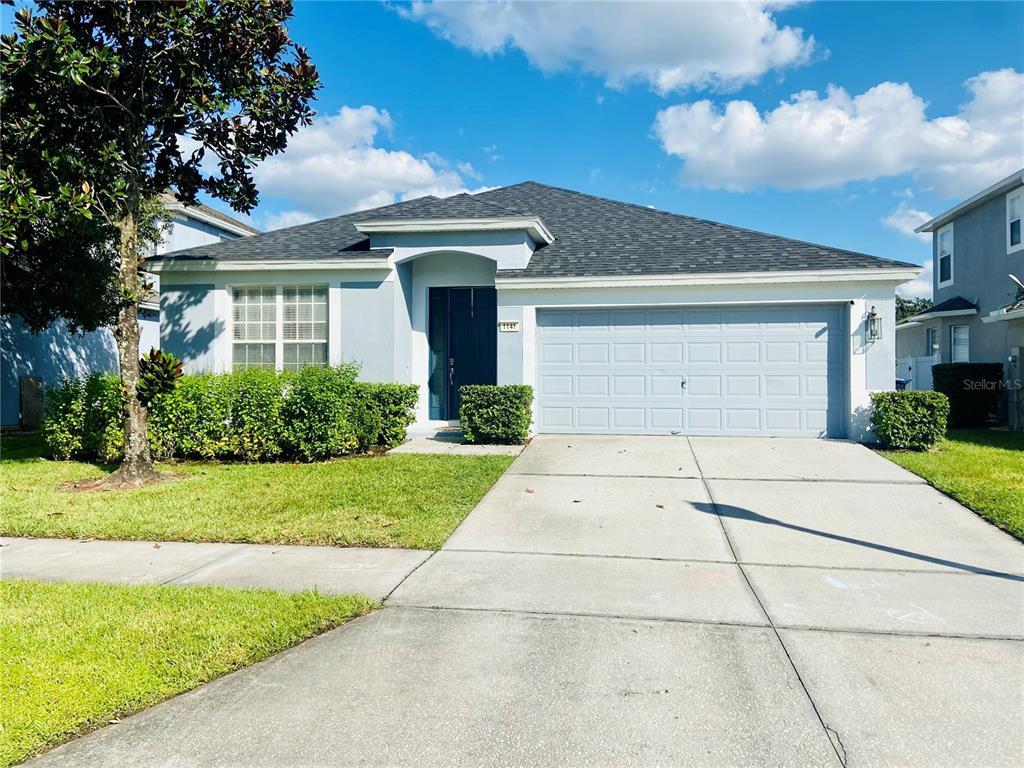 Light-blue single-story house with a wide garage, concrete driveway, and leafy tree at the left.