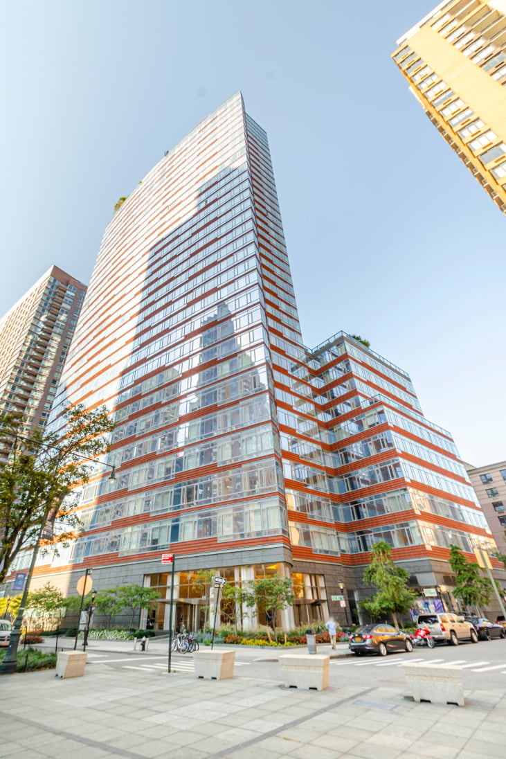 Residential high-rise with a glass facade, orange bands, and a paved plaza at the street corner.
