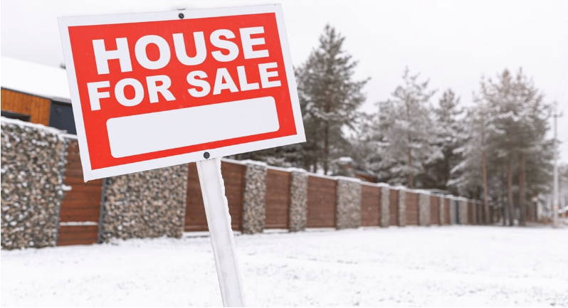 A red house-for-sale sign stands beside a snow-covered yard and long fence.