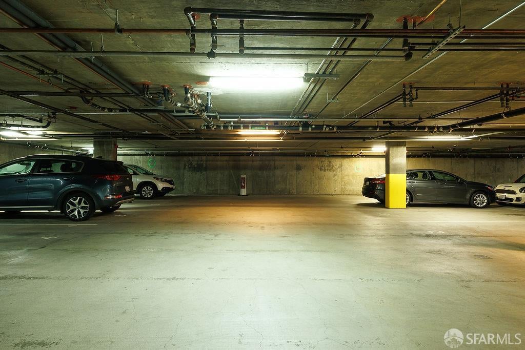 Underground parking garage with parked cars, exposed ceiling pipes, and a yellow-protected support column.
