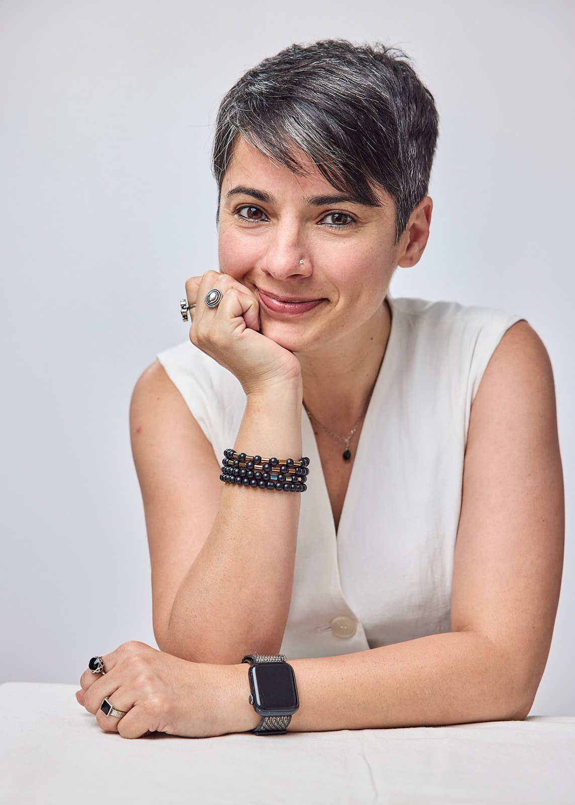 Rashi Malhotra smiling with her chin resting on one hand, wearing a white sleeveless top and dark bracelets.