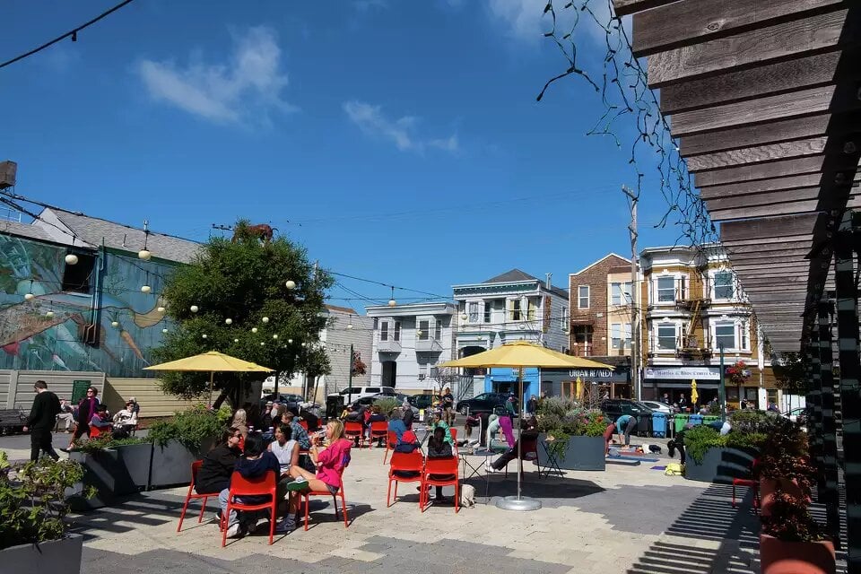 Community members gathering at Noe Valley Town Square during a farmers' market event, surrounded by local stalls.