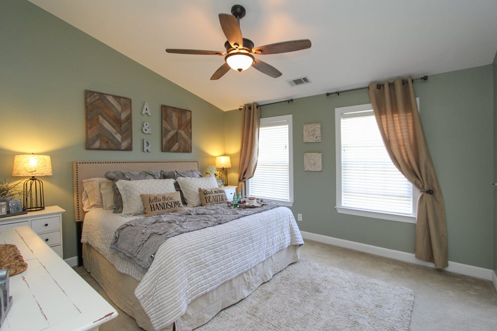 Furnished bedroom with a large bed, sage-green walls, two curtained windows, and a ceiling fan.