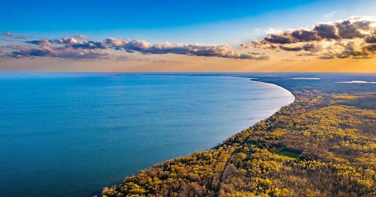 A broad lake curves beside an autumn-colored wooded shoreline at golden hour.